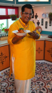 "Chef Ajay Chopra smiling while wearing his chef's uniform and holding a plate of beautifully presented gourmet food."