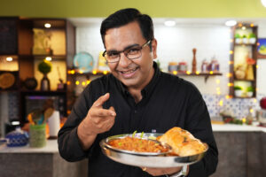 "Chef Ajay Chopra smiling while wearing his chef's uniform and holding a plate of beautifully presented gourmet food."